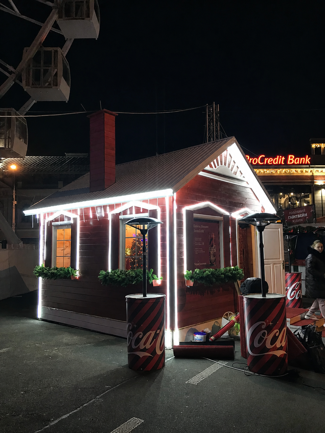 Außenansicht der Holzhütten-Aktivierung bei Nacht, mit Lichterketten und überdimensioniertem Coca-Cola-Fass