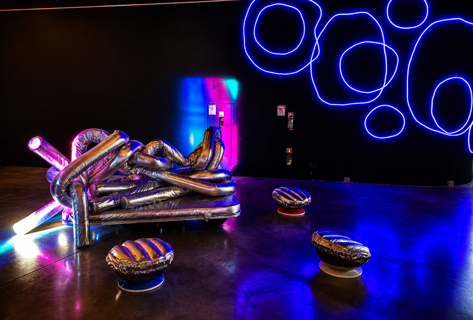Silberne aufblasbare Knoten-Sitzskulptur unter dem blauen Neon-Kreis-Mural